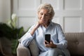 Happy older woman sitting on comfortable couch, looking at smartphone screen.