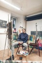 Happy painter sits on a chair near an easel with a canvas in his own studio, looks at the camera and smiles. Portrait of a Royalty Free Stock Photo