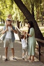 Happy parents with little kids at the park, cute daughter sitting on fathers shoulders, lovely mother lift in air joyful