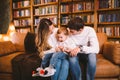 Happy parents playing with small child at Christmas time at home in living room on sofa near large cabinet with books and Royalty Free Stock Photo