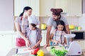 Happy parents stands behind their kids and smiling. They look at each other. Children cut vegetables separately.