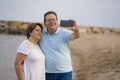 Happy pensioner woman and her husband taking romantic walk taking selfie - happy retired mature couple walking on the beach during