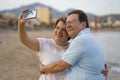 Happy pensioner woman and her husband taking romantic walk taking selfie - happy retired mature couple walking on the beach during