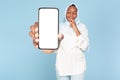 Happy pensive black muslim woman thinking, showing huge smartphone with blank screen, blue background, mockup