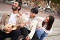 Happy people singing outdoors sitting in street. Group of friends having fun playing the ukulele. Royalty Free Stock Photo