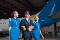 Happy pilot in uniform and aviator sunglasses walking together with two air stewardesses in blue uniform in front of big