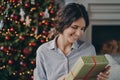 Happy pleased hispanic woman holding christmas gift box while sitting on sofa near xmas tree Royalty Free Stock Photo