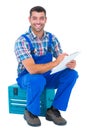 Happy plumber writing on clipboard while sitting on toolbox