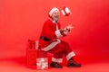 Happy positive gray bearded santa claus tossing up gift box and looking at camera with toothy smile, sitting near holidays