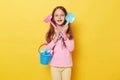 Happy positive little cute girl with dark hair holding beach sandbox toys rake and shovel isolated over yellow background having Royalty Free Stock Photo