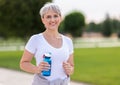 Happy positive mature woman with broad smile holding water bottle while doing sport in city park