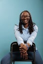 Happy positive smiling heartily young woman sitting on chair while looking at camera on blue background. Royalty Free Stock Photo