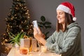 Happy positive woman wearing Santa cap looking at smartphone screen scrolling online doing shopping buying presents for Christmas Royalty Free Stock Photo