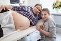 Happy pre teen daughter sitting near her pregnant mother with large belly in an ultrasonography room