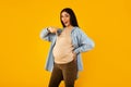 Happy pregnancy. Excited expectant woman pointing at her big belly and smiling at camera, yellow background