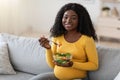 Happy pregnant black woman sitting on couch, eating fresh salad Royalty Free Stock Photo