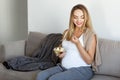 Happy pregnant blong young woman sitting on sofa eating bowl of fresh fruit  grey color of sofa and white top Royalty Free Stock Photo