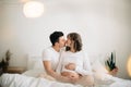 Happy pregnant couple relaxing on white bed and holding belly bump. Happy young husband kissing his smiling wife and hugging baby Royalty Free Stock Photo