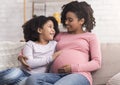 Happy pregnant mother and her cute afro daughter hugging on couch Royalty Free Stock Photo