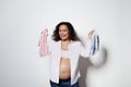 Happy pregnant woman with curly hair, holds pink and blue newborn clothes, expecting twins boy and girl, white backdrop Royalty Free Stock Photo