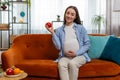 Happy pregnant woman eating fresh red apple fruit sitting on sofa healthy vitamin food motherhood Royalty Free Stock Photo