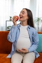 Happy pregnant woman eating fresh red apple fruit sitting on sofa healthy vitamin food motherhood Royalty Free Stock Photo