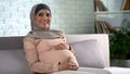 Happy pregnant woman in hijab touching belly gently, resting home, prenatal care