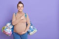 Happy pregnant woman holding bags with clothing for baby and herself to maternity house and pointing aside with index finger, Royalty Free Stock Photo