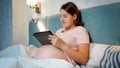 Happy pregnant woman in pajamas using tablet computer in bed before going to sleep at night Royalty Free Stock Photo