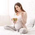 Happy pregnant young woman sitting and eating fruit salad on sofa at home Royalty Free Stock Photo