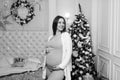 Happy pregnant woman sitting on bed and touching her belly at home over christmas tree Royalty Free Stock Photo