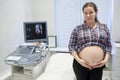 Happy pregnant woman standing in consulting room after ultrasonography near ultrasound scan with fetus 3d image on monitor screen