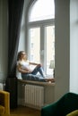 Happy pregnant woman touch bare tummy with hand sitting on windowsill in daylight. Home resting, relaxation near window.