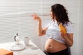 Happy pregnant young beautiful woman eating croissant and looking througt blinds during morning breakfast. Concept of Royalty Free Stock Photo