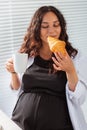 Happy pregnant young beautiful woman eating croissant during morning breakfast. Concept of pleasant morning and positive Royalty Free Stock Photo