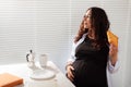 Happy pregnant young beautiful woman eating croissant during morning breakfast. Concept of pleasant morning and positive Royalty Free Stock Photo