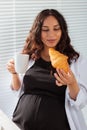 Happy pregnant young beautiful woman eating croissant during morning breakfast. Concept of pleasant morning and positive Royalty Free Stock Photo