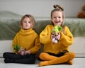 Happy pretty small girls sisters children in warm yellow knitted home sweaters sitting on floor and playing with dolls