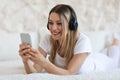 Happy blonde woman using cell phone and headset, bedroom interior Royalty Free Stock Photo