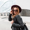 Happy pretty young hipster woman in stylish purple hat in trendy sunglasses in a black vintage jacket with a leather backpack Royalty Free Stock Photo