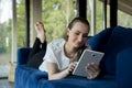 Happy pretty young woman in casual attire lies at home on a blue sofa and uses a tablet while browsing the Internet Royalty Free Stock Photo