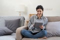 Happy pretty young woman in casual outfit sitting on sofa at home and using tablet while browsing internet Royalty Free Stock Photo