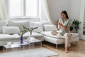 Happy pretty young woman in casual wear sitting on sofa at home and using smartphone while browsing internet Royalty Free Stock Photo