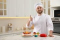 Happy professional confectioner with cakes showing thumb up gesture in kitchen