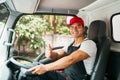 Happy professional truck driver with his assistant wearing a red cap thumb up, smiling, looking at the camera from a truck window Royalty Free Stock Photo