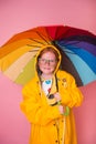 Happy red hair kid girl laughing with rainbow colored umbrella on pink background. Emotional freckled child in yellow Royalty Free Stock Photo