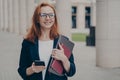 Happy red-haired business lady in official clothes standing outdoors, using mobile phone Royalty Free Stock Photo