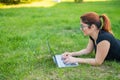 Happy red-haired girl in glasses lies on the lawn in the park and types on the laptop keyboard. Young caucasian female