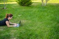 Happy red-haired girl in glasses lies on the lawn in the park and types on the laptop keyboard. Young caucasian female