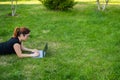Happy red-haired girl in glasses lies on the lawn in the park and types on the laptop keyboard. Young caucasian female